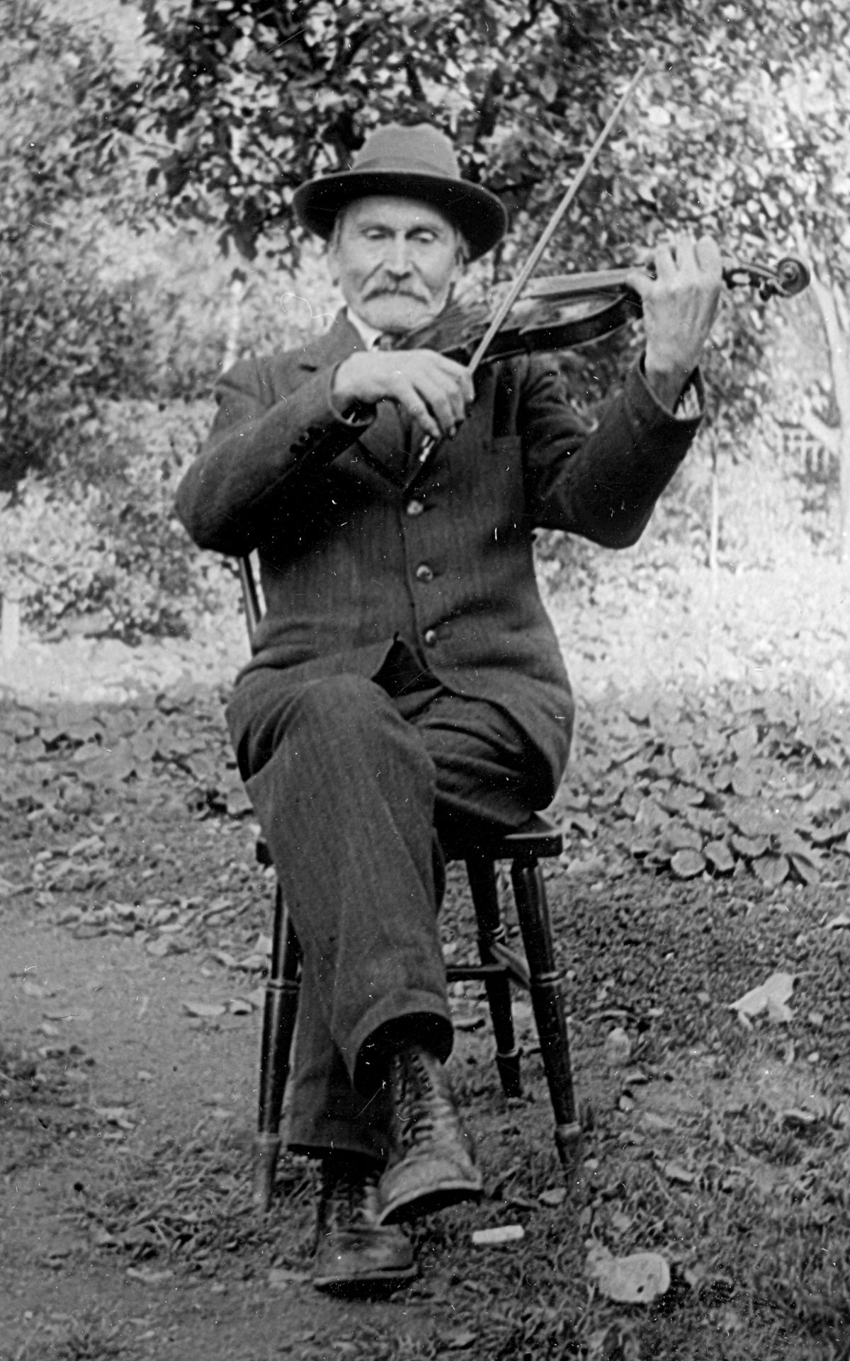 Bild på spelmannen Karl Herman Erlandsson som sitter och spelar fiol på en stol, utomhus.