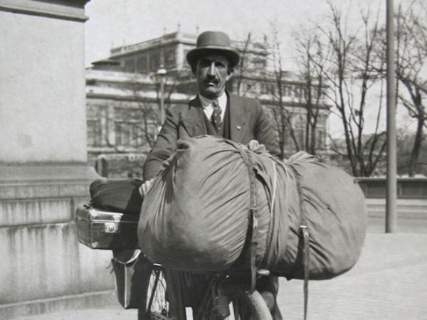 Svartvitt fotografi på en man som står med sin välpackade cykel. Mannen är klädd i kostym och bär hatt.