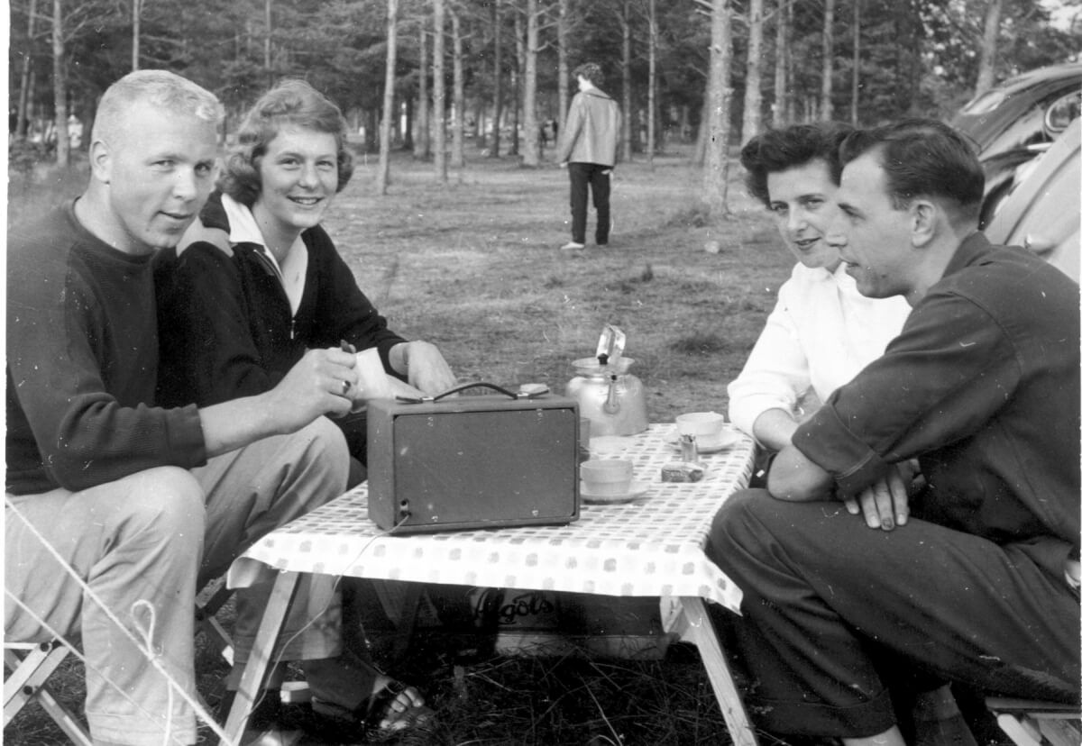 Fyra personer fikar runt ett fällbord och lyssnar på radio.